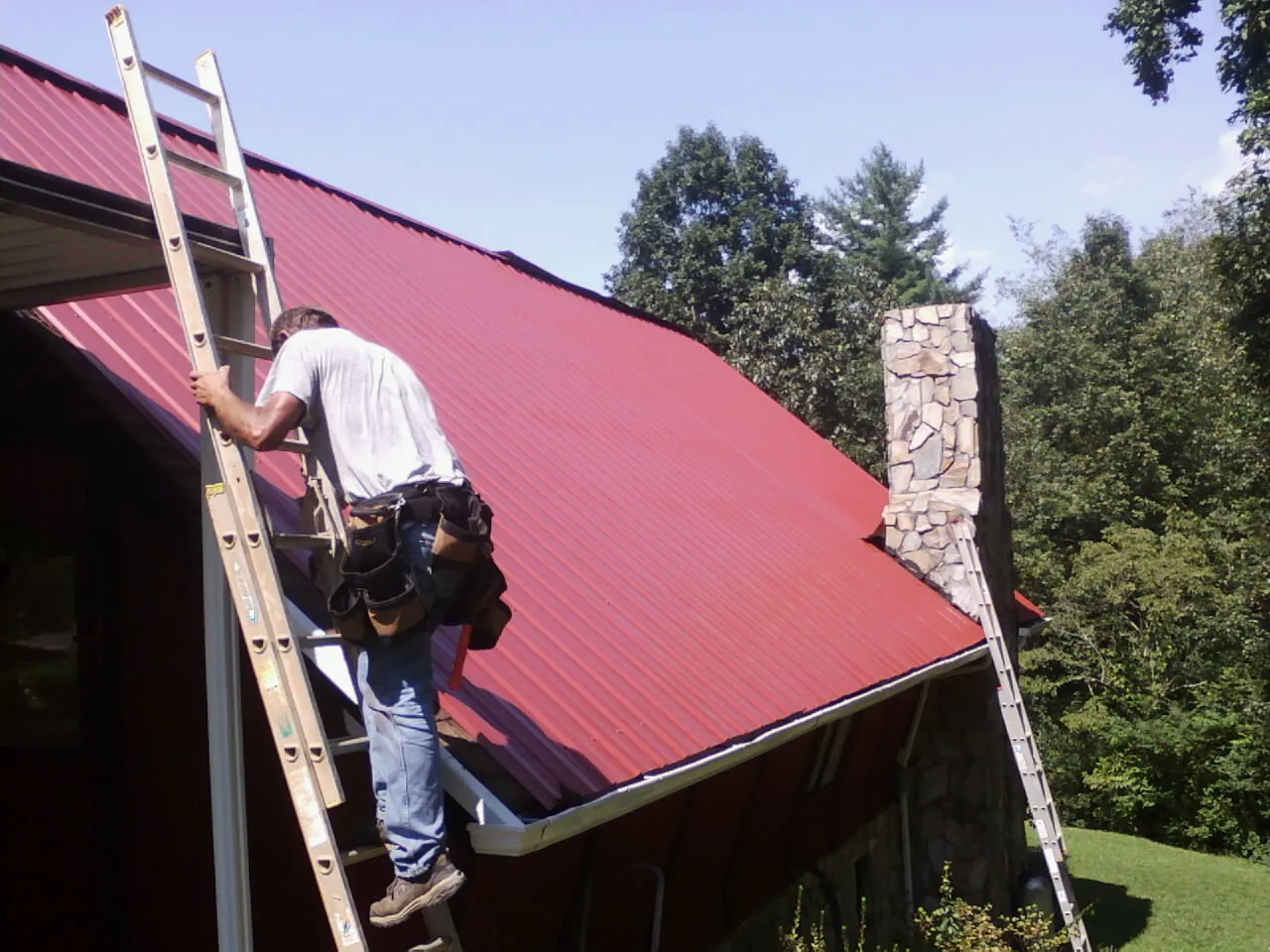 Roofer on a ladder beside a red metal roof and stone chimney — Soffit & Fascia Repair by Roofing Brothers in Sherrills Ford
